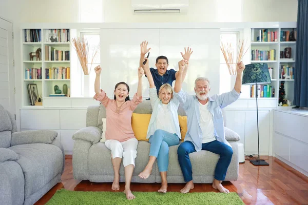 Szczęśliwy starszy wieloetnicznej rodziny siedzi na szarej kanapie w salonie i podnoszą ręce podczas oglądania piłki nożnej i ich zespół dostał zwycięzca z uśmiechniętą twarzą — Zdjęcie stockowe