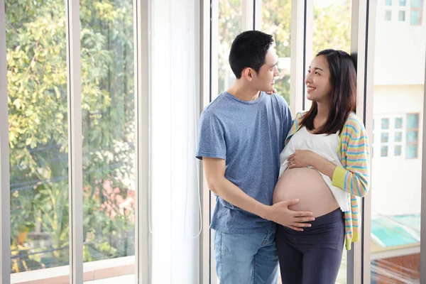 Junger asiatischer Ehemann steht und umarmt, die Augen ziehen sich zusammen mit jungen schwangeren Bäuchlein Frau mit glücklich lächelndem Gesicht und grünen Bäumen Hintergrund — Stockfoto