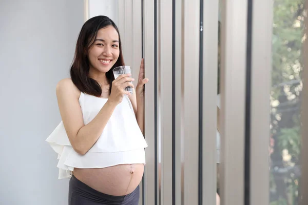 Joven asiático saludable embarazada vientre sonrisa mientras sostiene un vaso de agua con feliz cara y ojos contacto derecho copia espacio (salud cuidado concepto ) —  Fotos de Stock