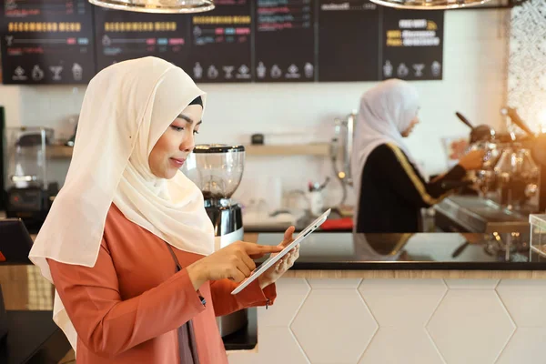 年轻的穆斯林女商人站在柜台，而使用平板电脑与年轻的咖啡师女孩工作模糊的背景 — 图库照片