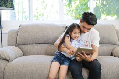 Mutlu Asyalı baba, kızına oturma odasındaki gri koltukta gülen ve gülen yüzlü tablet kullanmayı öğretiyor.)