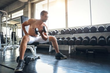 Bıyıklı yakışıklı beyaz adam gülümseyen yüzüyle spor salonunda halteri kaldırıyor.