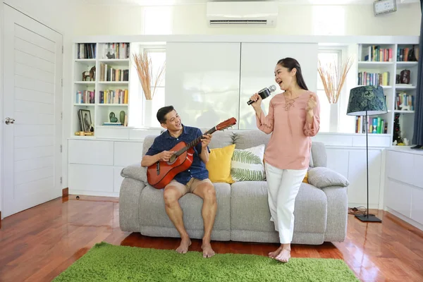 Pełny strzał szczęśliwego starszego azjatyckiego mężczyzny i kobiety, którzy śpiewają piosenkę i grają na gitarze na kanapie w salonie z szczęśliwą uśmiechniętą twarzą — Zdjęcie stockowe