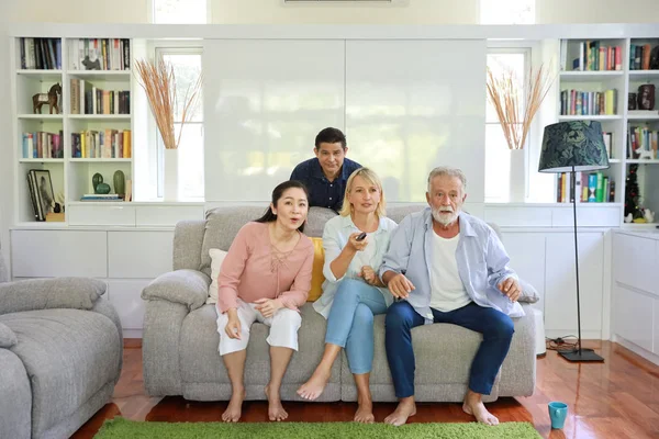 Pełny strzał z grupy szczęśliwego starszego wieloetniczna rodzina siedzi na szarej kanapie w salonie i oglądania piłki nożnej lub ulubiony kanał z ekscytującą twarz — Zdjęcie stockowe