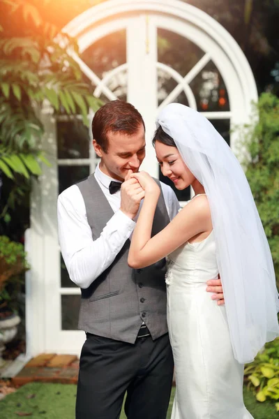 ハンサムな白人の新郎は、結婚式や結婚式前の写真のポーズの間に愛と笑顔でアジアの花嫁の手を保持し、キスしています — ストック写真