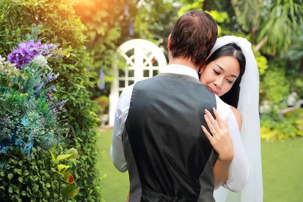 Unga asiatiska brud i vitt brudklänning är kramar sin man kaukasiska brudgum med kärlek medan nära hennes ögon och lycka under bröllopet i trädgården — Stockfoto