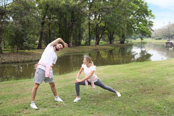 Imagen de pareja caucásica joven disfrutando en el ejercicio y estiramiento al aire libre con caras sonrientes y ojos en contacto con árboles verdes durante el verano en el parque — Foto de Stock