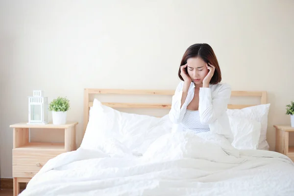 白いシャツを着た若い魅力的でセクシーなアジアの女性は、不幸な顔をした白い寝室のベッドで頭痛を起こしました. — ストック写真