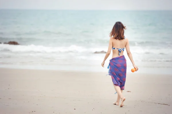 Visão traseira da menina bem torneada andando na praia tropical em maiô e segurando suco de laranja na natureza com céu azul — Fotografia de Stock