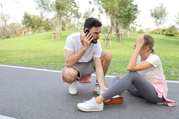 young girl fall down because of broken leg from too much exercise and young man is helping by using cell phone calling someone