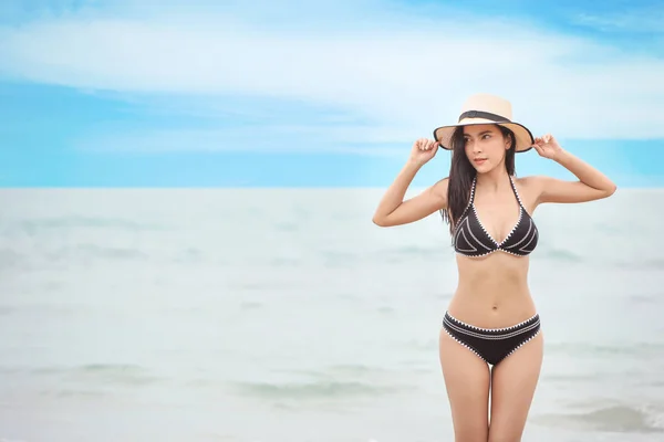Siyah bikinili ve beyaz şapkalı güzel ve seksi bir Asyalı kadının portresi kumlu sahilde duruyor mavi gökyüzü ve sol fotokopi alanı. — Stok fotoğraf