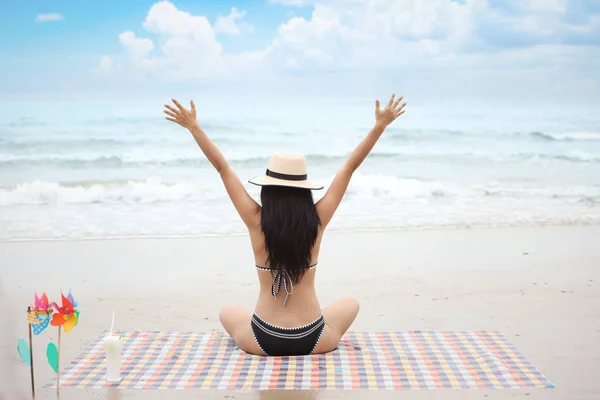 Volver ver retrato de chica joven sexy en bikini blanco y negro y sombrero en la playa tropical con las manos arriba mientras está sentado en tela de colores en la naturaleza con cielo azul . — Foto de Stock