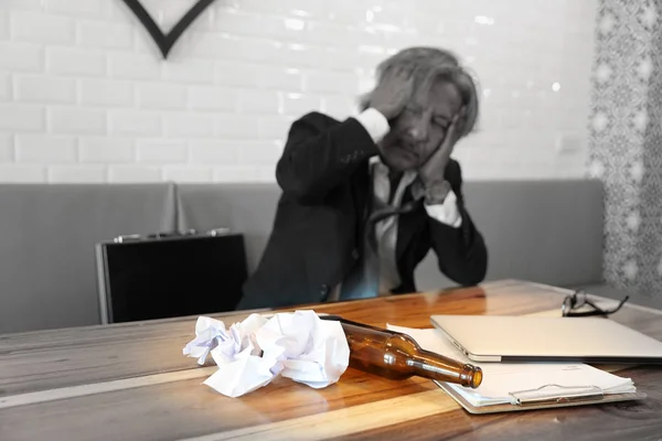 image of empty alcohol bottle on wood table with senior businessman who holding his head with stress because of failure in business blurred background