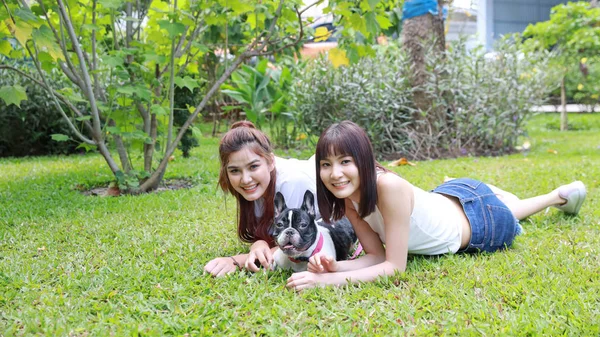 Hermosa mujer asiática dos jóvenes vistiendo camisa blanca que se encuentra en la hierba y jugando con su lindo perro con la cara feliz y sonriente en el jardín con árboles verdes. (concepto de amistad) —  Fotos de Stock