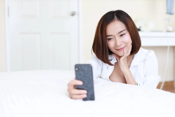 Primo piano bella donna asiatica, capelli corti in camicia bianca con videochiamata o autoritratto da cellulare su letto bianco in camera da letto con volto sorridente felice — Foto Stock