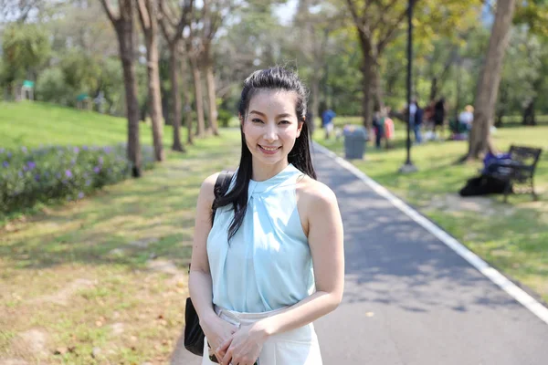 Portrait de belle femme asiatique portant chemise verte et sac à main noir debout dans le parc avec visage souriant et arbres verts — Photo