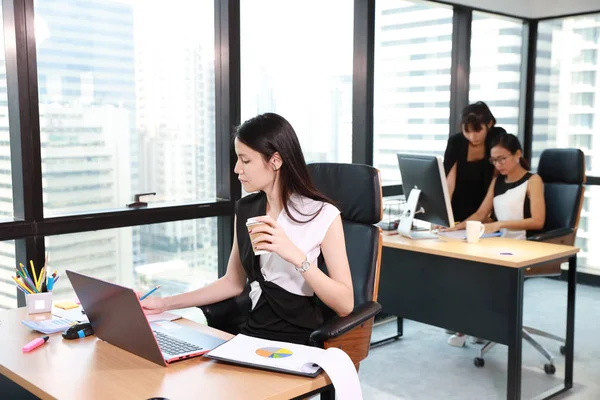 Kąt boczny widok azjatycki sekretarz kobieta lub biznesmen noszenie czarno-białej sukni pracy na laptopie z uśmiechniętą twarzą w nowoczesnym biurze z kolegami i widokiem na miasto tło — Zdjęcie stockowe