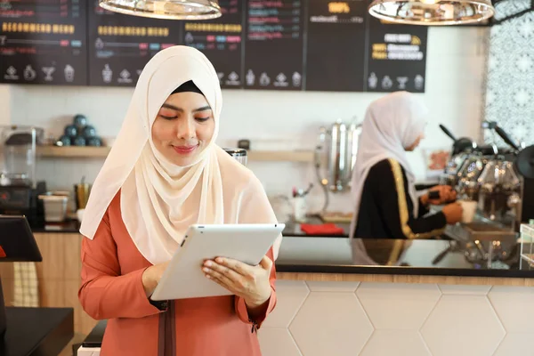 Jovem muçulmano empregado barista de pé e usando tablet no balcão de café com colega de trabalho desfocado fundo — Fotografia de Stock