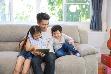 mutlu asyalı tek baba kızı ve oğlu kullanmak veya gülen ve gülümseyen yüzü ile oturma odasında gri kanepeüzerinde tablet oynamak öğretmek (teknoloji kavramı ile yaşam tarzı)