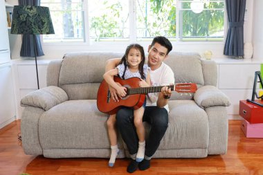 Mutlu Asyalı baba kızına oturma odasındaki gri koltukta gülen yüzlü akustik gitar çalmayı öğretiyor.)