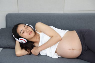 uykulu genç anne, hamile asyalı kadın oturma odasında kulaklık ile kanepede yatan 
