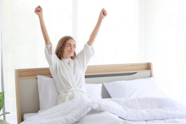 Neşeli ve sağlıklı beyaz pijama giyen genç bir kadın sabah uyanıp yatak odasında dinleniyor ve gülümsüyor.