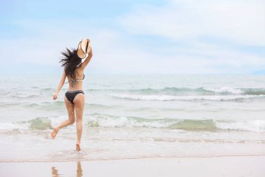 arka görüş genç asyalı güzel ve seksi kadın, siyah bikini uzun saçlı, kumlu plajda mavi gökyüzü ile beyaz şapka tutarken havada atlama (tatil tatil konsepti)