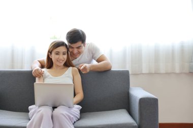 Genç Asyalı hamile kadın kanepeye oturmuş bilgisayardan alışveriş yapıyor ve beyaz bir koca ekranda gülümseyen bir yüzü işaret ediyor.