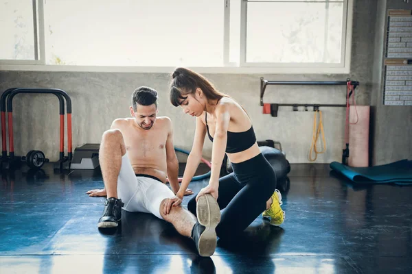 Mladá běloška žena pomáhá fitness muž, který nosí sportovní oblek a zranil kotník z příliš mnoho cvičení v tělocvičně — Stock fotografie