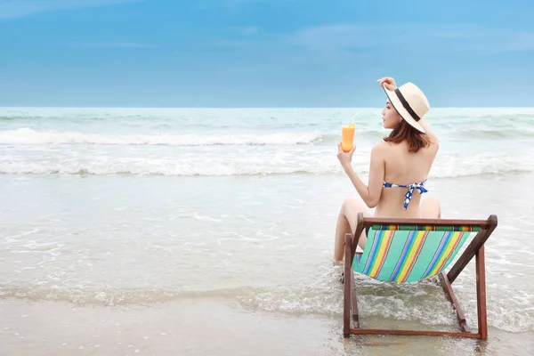 Портрет сексуальной азиатской молодой девушки в бикини и шляпе, сидящей на шезлонге на тропическом пляже и держащей апельсиновый сок с красивым лицом на отдыхе в счастливое время лета — стоковое фото