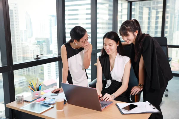 three asian colleagues businesswomen whispering or gossiping someone from internet with laughing while working at the office with computer