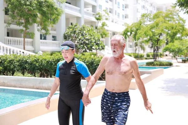 Feliz anciano caucásico marido y anciano asiático esposa cogido de la mano mientras camina a la piscina en el hotel en camino caliente y clima caliente en día soleado al aire libre — Foto de Stock