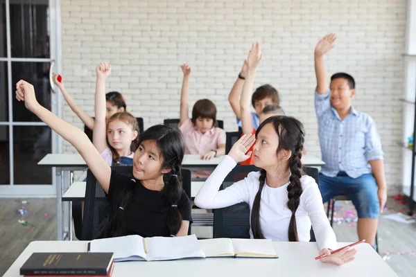 Söta små elever höjer händerna i klassrummet med glatt leende ansikte (utbildningskoncept) — Stockfoto