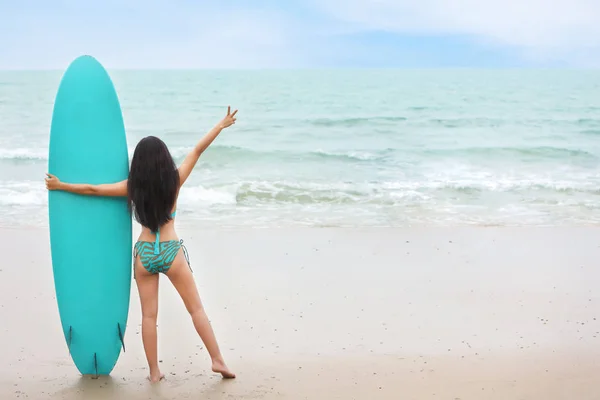 Vissza kilátás szép és szexi szörfös lány gazdaság fedélzeten a trópusi tengeren zöld fürdőruhát a nyáron a tengerparton, míg ő kezét, és nem győzelem szimbóluma. (vízisport vagy nyaralás ünnep koncepció) — Stock Fotó