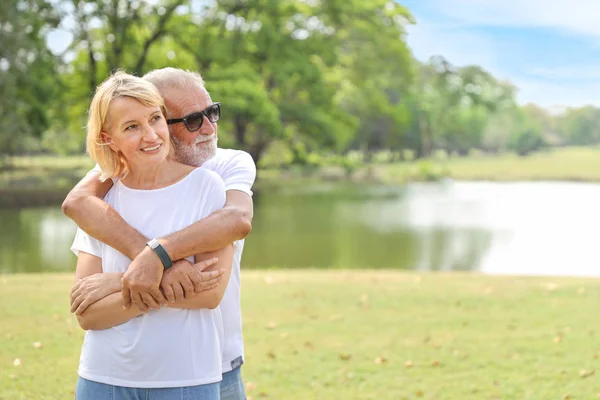 elderly couple with white shirt, blue jean and sun glasses standing and embracing in park during summer time on wedding anniversary day