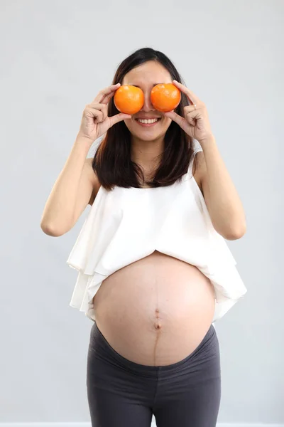 Longitud completa disparo joven asiático embarazada vientre en blanco vestido con feliz sonriente cara celebración de naranjas, mientras que de pie sobre blanco pared fondo —  Fotos de Stock