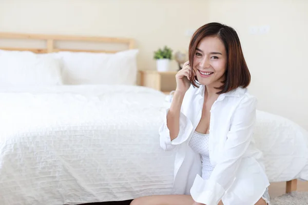 Vista lateral de adulto freelance mujer asiática en camisa blanca trabajando en la computadora y el teléfono celular en el dormitorio con la cara sonriente feliz —  Fotos de Stock