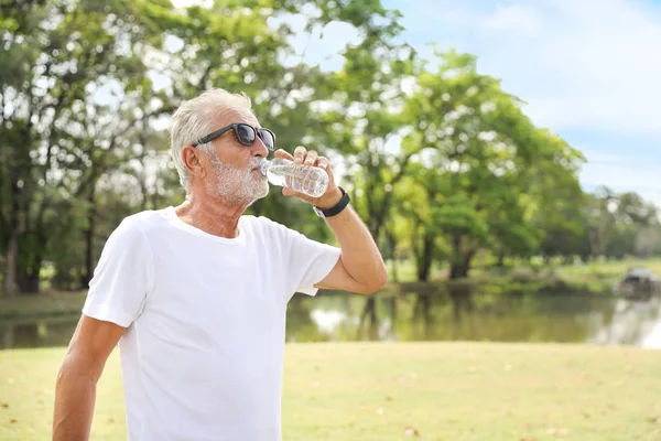 beyaz gömlek giyen yaşlı beyaz adam ve güneş gözlüğü tutan ve gülümseyerek parkta saf su şişesi içme. tatillerde ferahlatıcı 