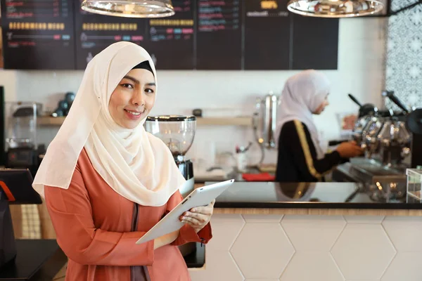 Jovem muçulmano empregado barista de pé e usando tablet no balcão de café com colega de trabalho desfocado fundo — Fotografia de Stock