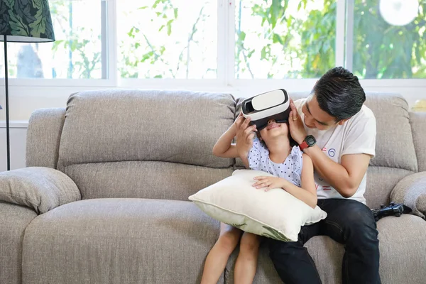 Happy Asian family, father teaching daughter to play virtual reality goggles on the sofa in the living room with happy smiling face (relaxation and technology concept)