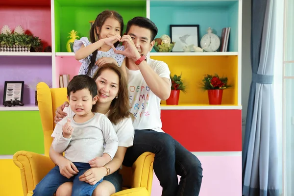 Happy asian family father, mother, child daughter and son sitting on sofa in colorful modern living room doing heart symbol with relaxation and smiling face