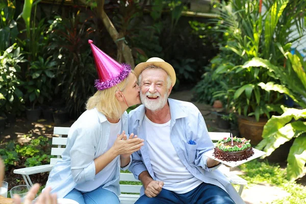 Caucasian elder wife kissing her caucasian husband with happy smiling face on anniversary day while sitting in backyard having breakfast and her husband holding birthday cake