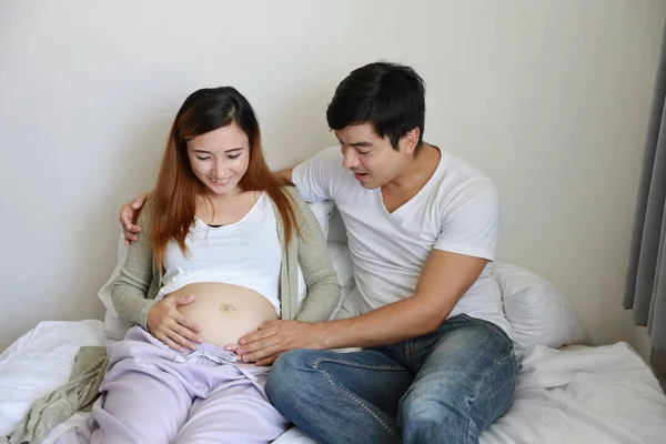 Bello caucasico marito toccare la sua asiatico incinta pancia moglie con felice sorriso faccia e amore su letto in bedroo — Foto Stock