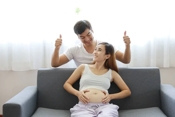 Junge kaukasische Ehemann hebt seine Hände und Daumen nach oben vor Glück und fröhlich zu seiner jungen asiatischen schwangeren Bauch Frau, während sie auf dem Sofa im Wohnzimmer sitzen — Stockfoto