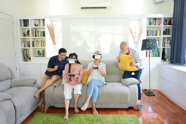 Happy elder multiethnic family sitting on grey sofa in living room and playing game from virtual reality goggles with smiling face