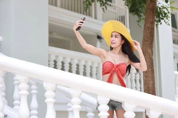 穿着橙色比基尼、头戴漂亮帽子的迷人而性感的亚洲女孩用手机拍照，脸上挂着快乐的笑容 — 图库照片