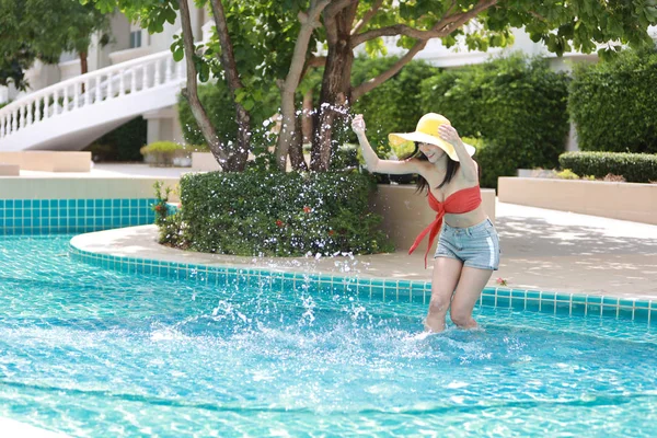 Atractiva y sexy mujer asiática con sombrero, pelos largos en bikini naranja jugando al agua en la piscina durante el día soleado con felicidad y la cara risueña — Foto de Stock