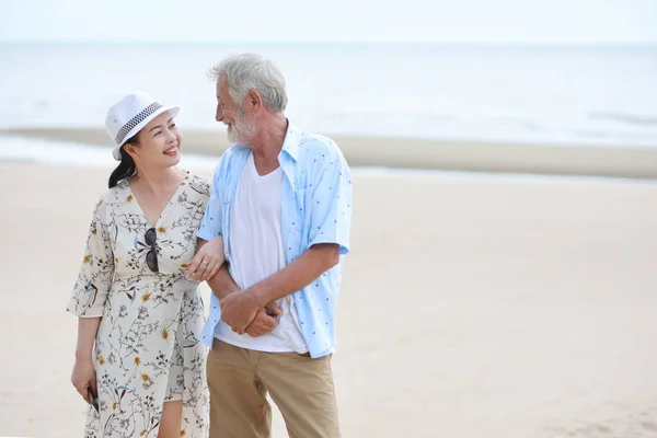 older couple caucasian husband and asian wife holding hand and resting and standing on sandy beach after retirement in resort during the summer with right copy space