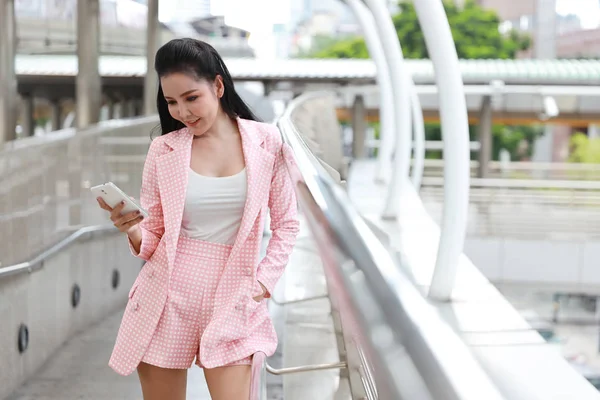 Frente vista bem sucedida asiática empresária em rosa vestido casual que de pé no caminho a pé no centro da cidade, ela usando telefone celular com feliz rosto sorridente com fundo da cidade — Fotografia de Stock