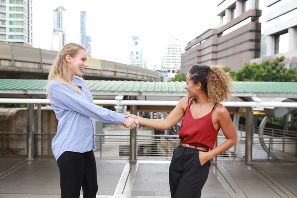 half shot of two caucasian businesswomen shaking hands together meaning of teamwork while walking on walk way with city and skytrain background